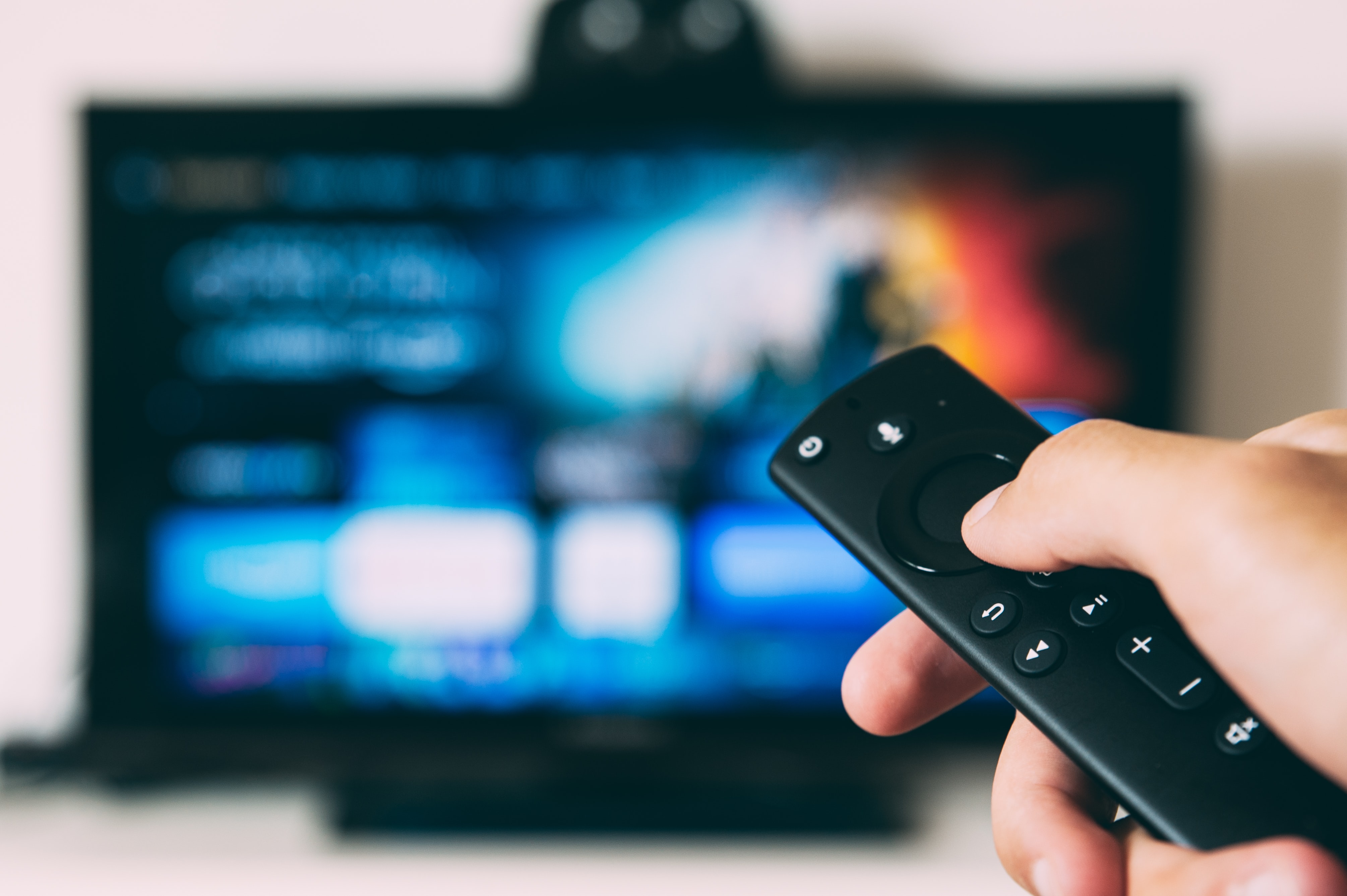 A hand holds up a remote in front of a large television screen, blurred in the background.